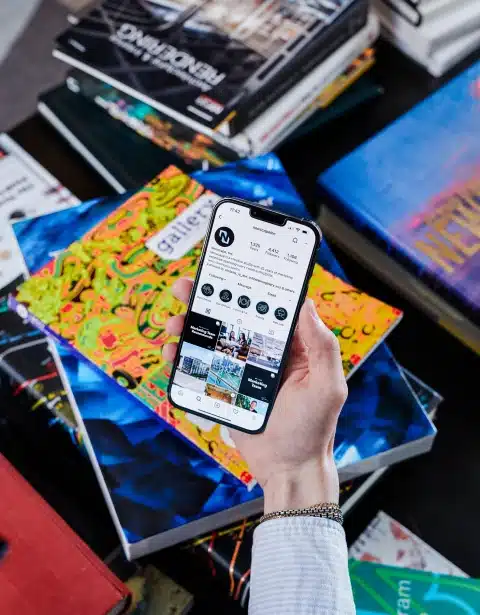 Hand holding smartphone displaying Instagram feed, surrounded by design books and colorful covers