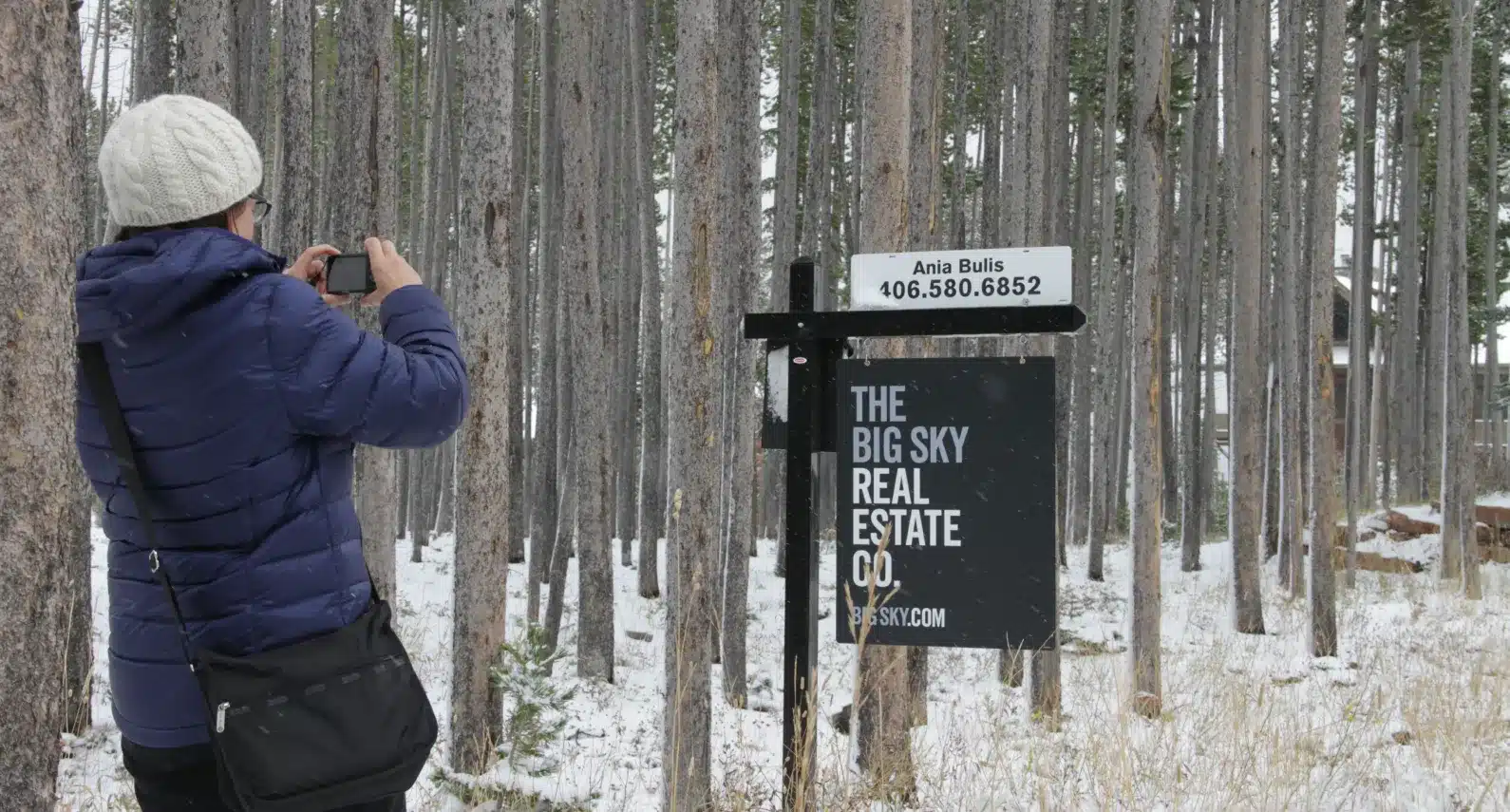 A woman in a winter coat photographs a real estate sign in a snowy pine forest