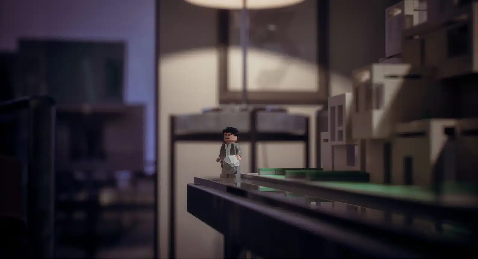 Close-up of LEGO figure posed beside architectural model, with blurred background of warm apartment lighting